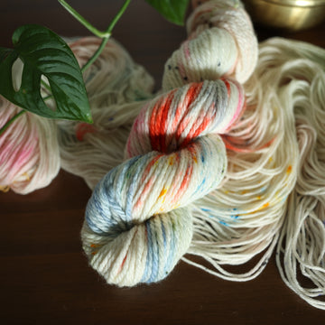 bulky white yarn with mixed colors of light pink, purple, blue, and red/orange with speckles, on top of an unwound version of that same yarn. all on top of a dark wooden table with a couple of leaves of a plant overhead in a gold pot.