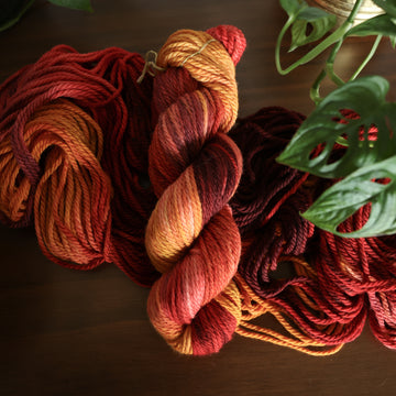 Bulky yarn with red orange and yellow colors, on top of an unwound version of that same yarn. all on top of a dark wooden table with a couple of leaves of a plant overhead in a gold pot.