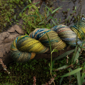 A Bulky yarn skein colored with green, yellow and grey colors, with blue speckles, on top of a tree limb that is hovering over a pond, with different mossy plants around the pond, and around the yarn.