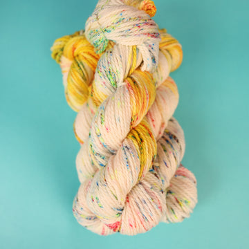A light cream colored Bulky yarn with different colored speckles all over and banana colored yellow at the top of the skein on top of a light blue background.