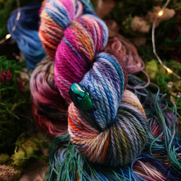 Bulky yarn colored with a hot pink, light blue, dark blue/purple, with some green and orange/brown colors, on top of the same yarn but unwound, with moss underneath and some fairy lights around the moss.