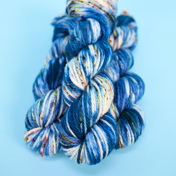 Bulky yarn skein colored with different shades of blue and white with multicolored speckles on the portion of the yarn that Is white. all on top of a light blue background.