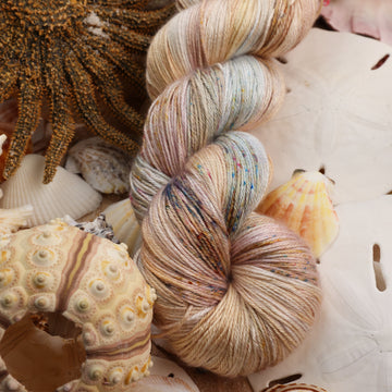 Silk yarn that is colored with different light browns and creme colors along with some blues and speckles of blue brown and pink. This yarn is on top of a bunch of different types  of light colored shells and sea stars and sand dollars.