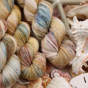 This yarn is colored with different light browns and creme colors along with some blues and speckles of blue brown and pink. This yarn is on top of a bunch of different types  of light colored shells and sea stars and sand dollars.