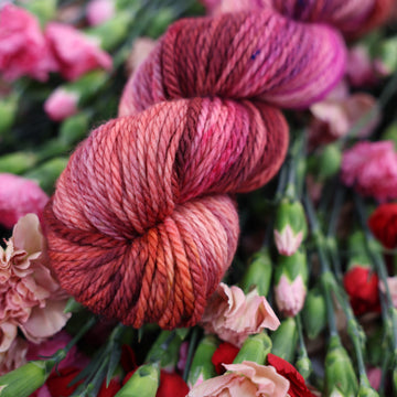 A Bulky skein of yarn colored with various amounts of lavenders, reds, pinks, fuschia, pale yellows and purples. This skein is sitting on top of colorful flowers.