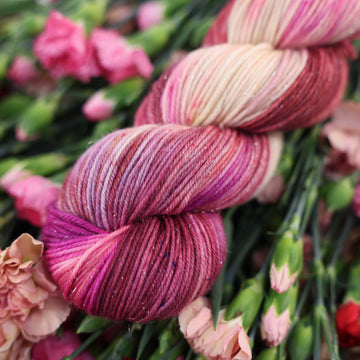A DK Sparkle skein of yarn colored with various amounts of lavenders, reds, pinks, fuschia, pale yellows and purples. This skein is sitting on top of colorful flowers.