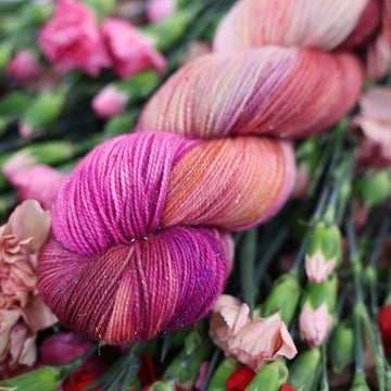 A Sparkle fingering skein of yarn colored with various amounts of lavenders, reds, pinks, fuschia, pale yellows and purples. This skein is sitting on top of colorful flowers.