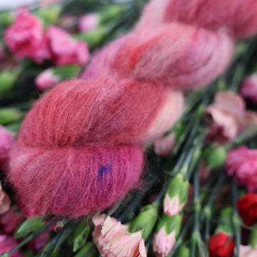 A Carnation Suri skein of yarn colored with various amounts of lavenders, reds, pinks, fuschia, pale yellows and purples. This skein is sitting on top of colorful flowers.