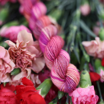 A DK mini skein of yarn colored with various amounts of lavenders, reds, pinks, fuschia, pale yellows and purples. This skein is sitting on top of colorful flowers.