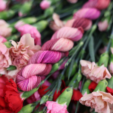 A Mini Fingering skein of yarn colored with various amounts of lavenders, reds, pinks, fuschia, pale yellows and purples. This skein is sitting on top of colorful flowers.