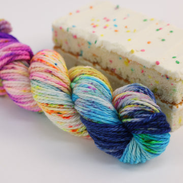 A Bulky skein of yarn colored bright neon blues, oranges, green, and hot pinks and blues and spearmint green. Next to it is a confetti cake bar, all sitting on a white table.