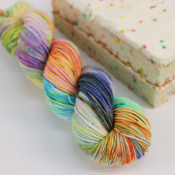 A DK skein of yarn colored bright neon blues, oranges, green, and hot pinks and blues and spearmint green. Next to it is a confetti cake bar, all sitting on a white table.