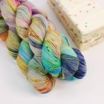 A sock skein of yarn colored bright neon blues, oranges, green, and hot pinks and blues and spearmint green. Next to it is a confetti cake bar, all sitting on a white table.