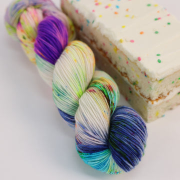 A sport skein of yarn colored bright neon blues, oranges, green, and hot pinks and blues and spearmint green. Next to it is a confetti cake bar, all sitting on a white table.