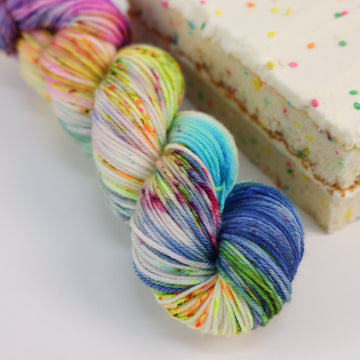 A worsted skein of yarn colored bright neon blues, oranges, green, and hot pinks and blues and spearmint green. Next to it is a confetti cake bar, all sitting on a white table.