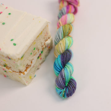 A DK Mini skein of yarn colored bright neon blues, oranges, green, and hot pinks and blues and spearmint green. Next to it is a confetti cake bar, all sitting on a white table.