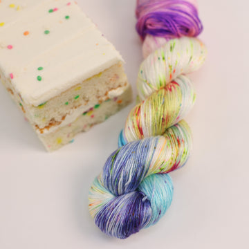 A silk skein of yarn colored bright neon blues, oranges, green, and hot pinks and blues and spearmint green. Next to it is a confetti cake bar, all sitting on a white table.