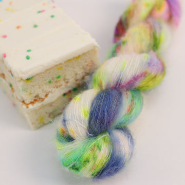 A suri skein of yarn colored bright neon blues, oranges, green, and hot pinks and blues and spearmint green. Next to it is a confetti cake bar, all sitting on a white table.