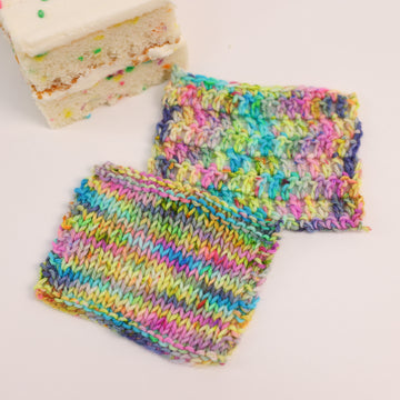 A two knit and crochet swatches colored bright neon blues, oranges, green, and hot pinks and blues and spearmint green. Next to it is a confetti cake bar, all sitting on a white table.