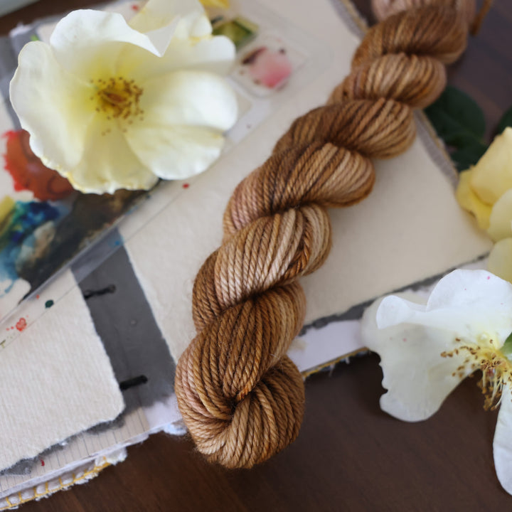DK mini yarn that is colored with different shades of brown and some dark brown speckles. This is on top of a brown table with a watercolor paint pallet on top of some canvas paper and some white and yellow flowers.