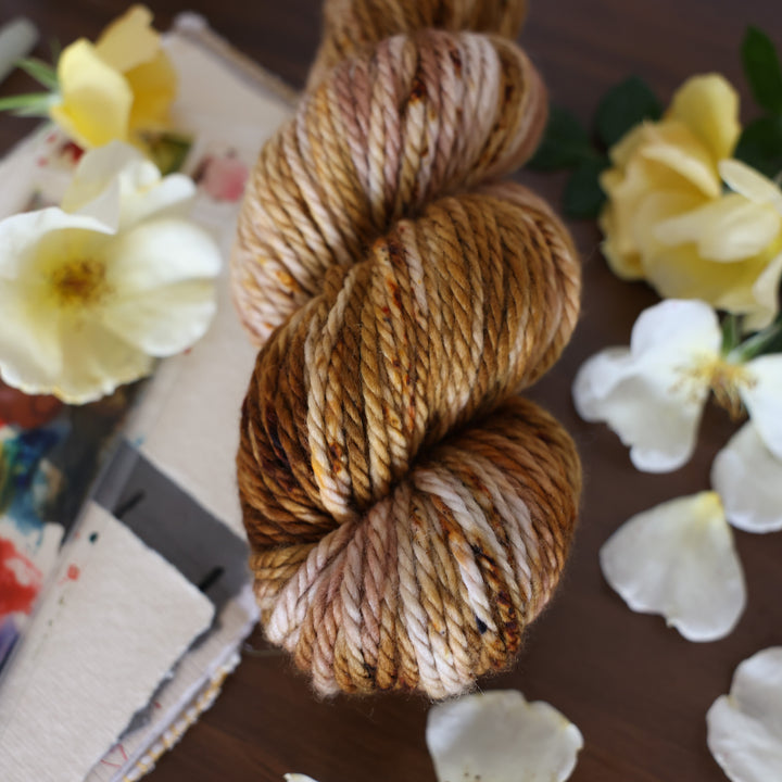 Bulky yarn that is colored with different shades of brown and some dark brown speckles. This is on top of a brown table with a watercolor paint pallet on top of some canvas paper and some white and yellow flowers.