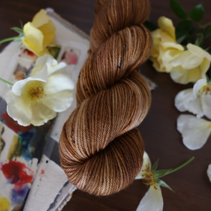 DK yarn that is colored with different shades of brown and some dark brown speckles. This is on top of a brown table with a watercolor paint pallet on top of some canvas paper and some white and yellow flowers.