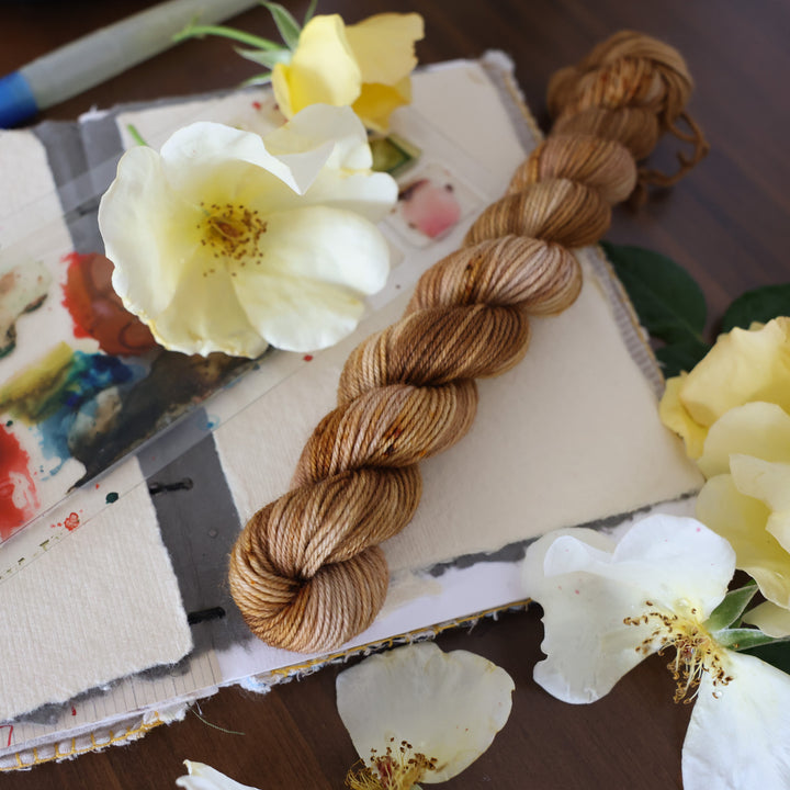 Fingering mini yarn that is colored with different shades of brown and some dark brown speckles. This is on top of a brown table with a watercolor paint pallet on top of some canvas paper and some white and yellow flowers.