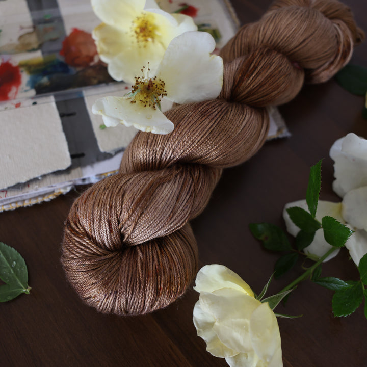 Silk yarn that is colored with different shades of brown and some dark brown speckles. This is on top of a brown table with a watercolor paint pallet on top of some canvas paper and some white and yellow flowers.
