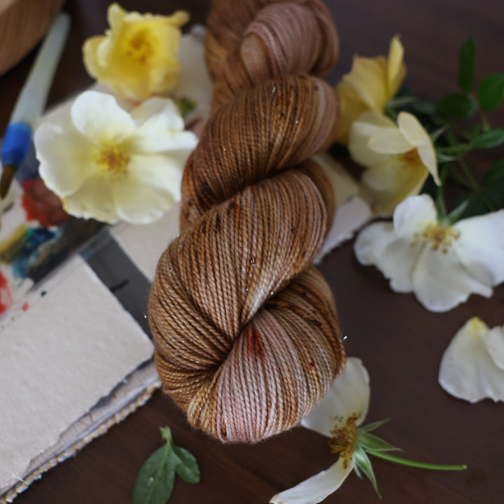 Sparkle yarn that is colored with different shades of brown and some dark brown speckles. This is on top of a brown table with a watercolor paint pallet on top of some canvas paper and some white and yellow flowers.