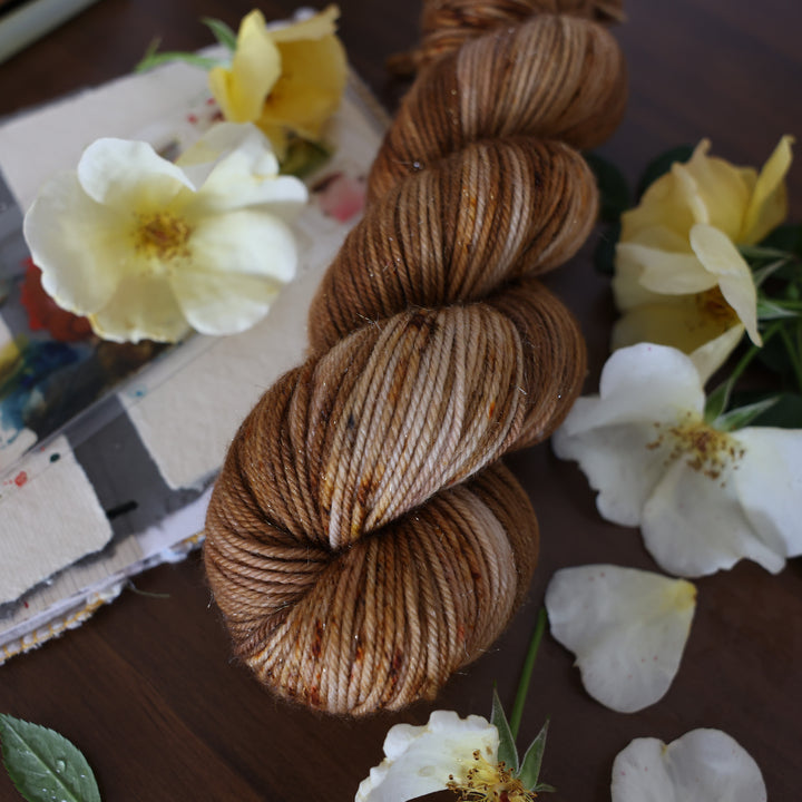 Sparkle DK yarn that is colored with different shades of brown and some dark brown speckles. This is on top of a brown table with a watercolor paint pallet on top of some canvas paper and some white and yellow flowers.