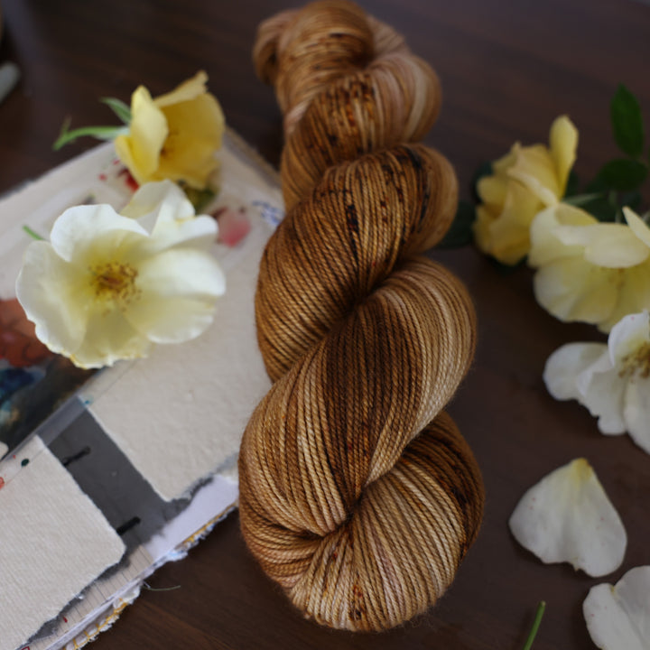 Sport yarn that is colored with different shades of brown and some dark brown speckles. This is on top of a brown table with a watercolor paint pallet on top of some canvas paper and some white and yellow flowers.