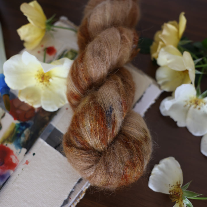 Suri yarn that is colored with different shades of brown and some dark brown speckles. This is on top of a brown table with a watercolor paint pallet on top of some canvas paper and some white and yellow flowers.
