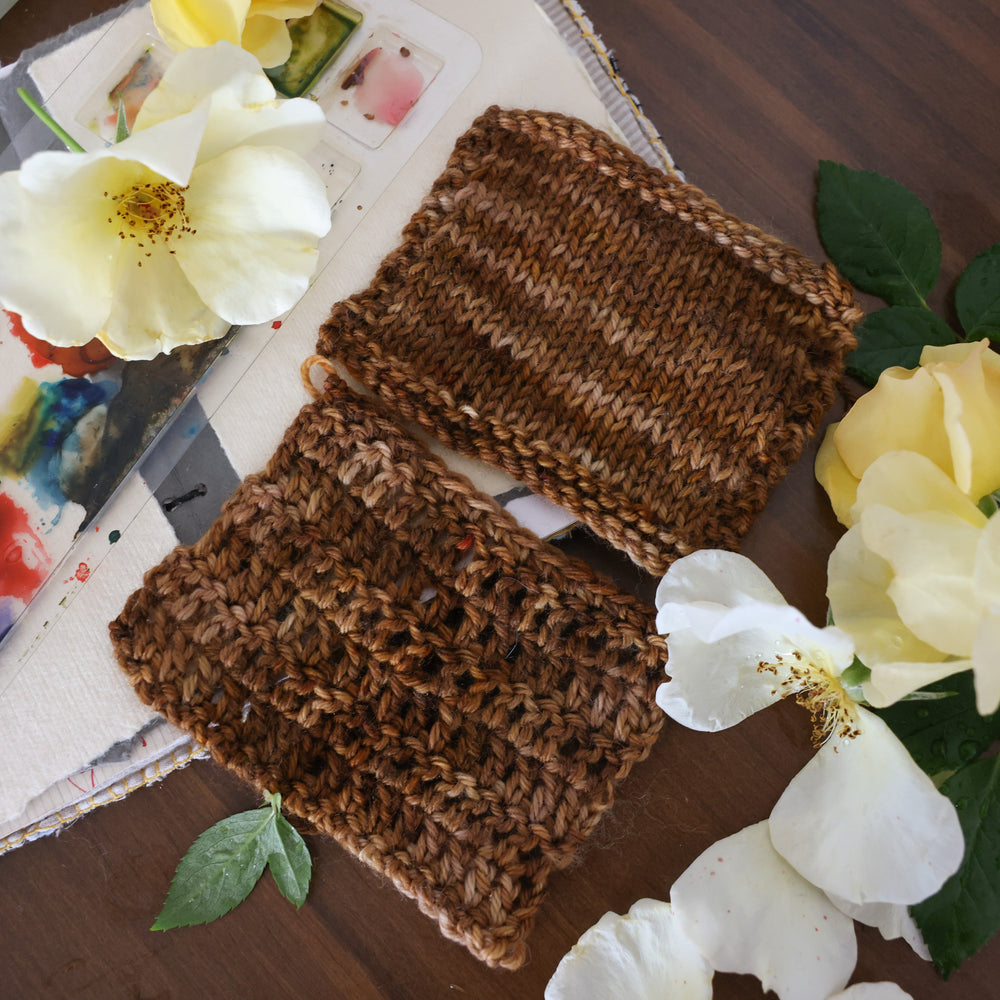 Crochet and knit swatches colored with different shades of brown. This is on top of a brown table with a watercolor paint pallet on top of some canvas paper and some white and yellow flowers.