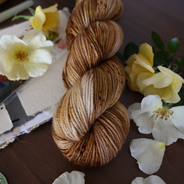 Worsted yarn that is colored with different shades of brown and some dark brown speckles. This is on top of a brown table with a watercolor paint pallet on top of some canvas paper and some white and yellow flowers.