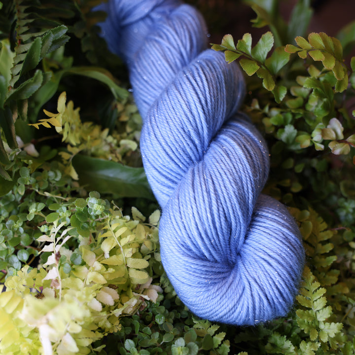 Sparkle DK yarn colored a light blue. This skein is on top of a bed of ferns and plants.