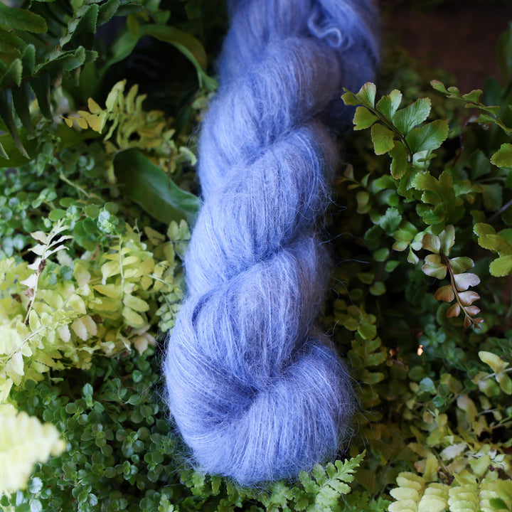Suri alpaca yarn colored a light blue. This skein is on top of a bed of ferns and plants.