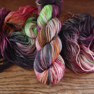 An unwound yarn skein that that colored with different shades of darker purples, light oranges, light pinks, and some lime greens mixed in, along with some other colors, along with a wound version of the same DK yarn on top, all on top of a wooden table.