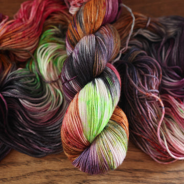 An unwound yarn skein that that colored with different shades of darker purples, light oranges, light pinks, and some lime greens mixed in, along with some other colors, along with a wound version of the same fingering yarn on top, all on top of a wooden table.