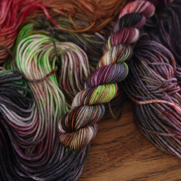 An unwound yarn skein that that colored with different shades of darker purples, light oranges, light pinks, and some lime greens mixed in, along with some other colors, along with a wound version of the same fingering mini yarn on top, all on top of a wooden table.