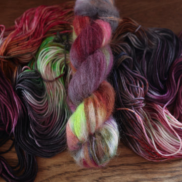 An unwound yarn skein that that colored with different shades of darker purples, light oranges, light pinks, and some lime greens mixed in, along with some other colors, along with a wound version of the same suri yarn on top, all on top of a wooden table.