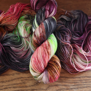 An unwound yarn skein that that colored with different shades of darker purples, light oranges, light pinks, and some lime greens mixed in, along with some other colors, along with a wound version of the same Sport yarn on top, all on top of a wooden table.