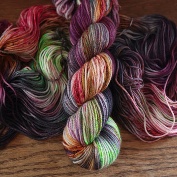An unwound yarn skein that that colored with different shades of darker purples, light oranges, light pinks, and some lime greens mixed in, along with some other colors, along with a wound version of the same Worsted yarn on top, all on top of a wooden table.