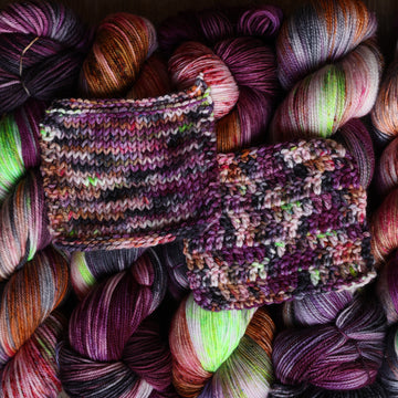 Yarn skeins stacked next to each other that are colored with different shades of darker purples, light oranges, dark and light pinks, and some lime greens mixed in, along with some other colors, with a knitted swatch and a crochet swatch on top.