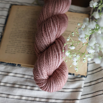 A Bulky skein of yarn colored a lovely pale mauve tonal-solid, sitting on top of a book, off to the corner there is white flowers hovering over the book. All is sitting on a black and white striped tablecloth. 