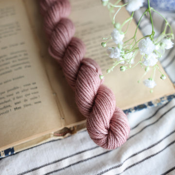 A DK Mini skein of yarn colored a lovely pale mauve tonal-solid, sitting on top of a book, off to the corner there is white flowers hovering over the book. All is sitting on a black and white striped tablecloth. 
