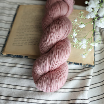 A fingering skein of yarn colored a lovely pale mauve tonal-solid, sitting on top of a book, off to the corner there is white flowers hovering over the book. All is sitting on a black and white striped tablecloth. 