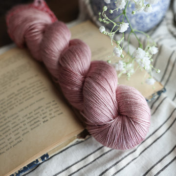 A Silk skein of yarn colored a lovely pale mauve tonal-solid, sitting on top of a book, off to the corner there is a blue and white vase with white flowers in it, the white flowers are hovering over the book. All is sitting on a black and white striped tablecloth. 