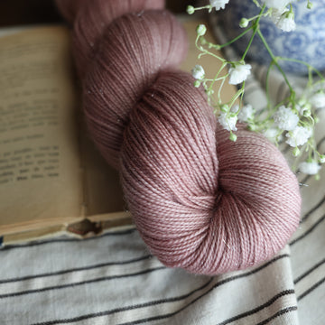 A Sparkle fingering skein of yarn colored a lovely pale mauve tonal-solid, sitting on top of a book, off to the corner there is a blue and white vase with white flowers in it, the white flowers are hovering over the book. All is sitting on a black and white striped tablecloth. 