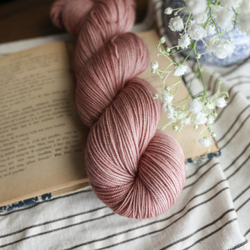 A sport skein of yarn colored a lovely pale mauve tonal-solid, sitting on top of a book, off to the corner there is a blue and white vase with white flowers in it, the white flowers are hovering over the book. All is sitting on a black and white striped tablecloth. 