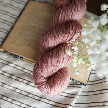 A Worsted skein of yarn colored a lovely pale mauve tonal-solid, sitting on top of a book, off to the corner there is white flowers hovering over the book. All is sitting on a black and white striped tablecloth. 